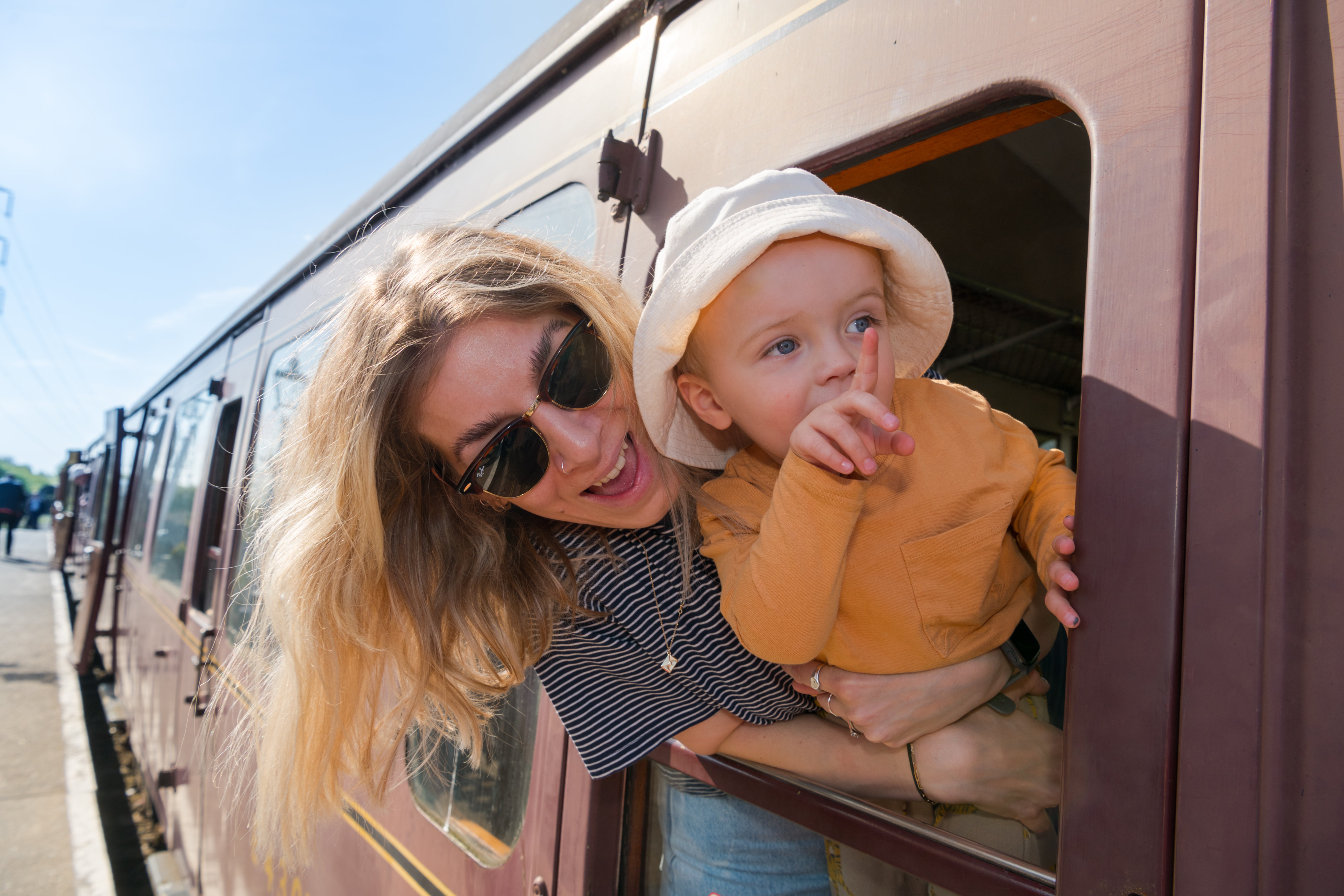 A woman and toddler stick their heads out of a train carriage window. The toddler is pointing at something off camera and they both look excited A woman and toddler stick their heads out of a train carriage window. The toddler is pointing at something off camera and they both look excited