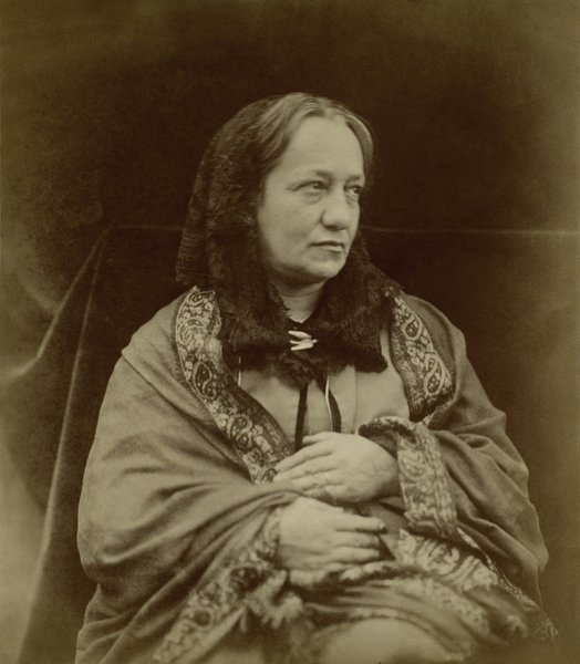 A sepia photograph of a seated woman. She is wearing a dark hood and a shawl with a paisley design border.