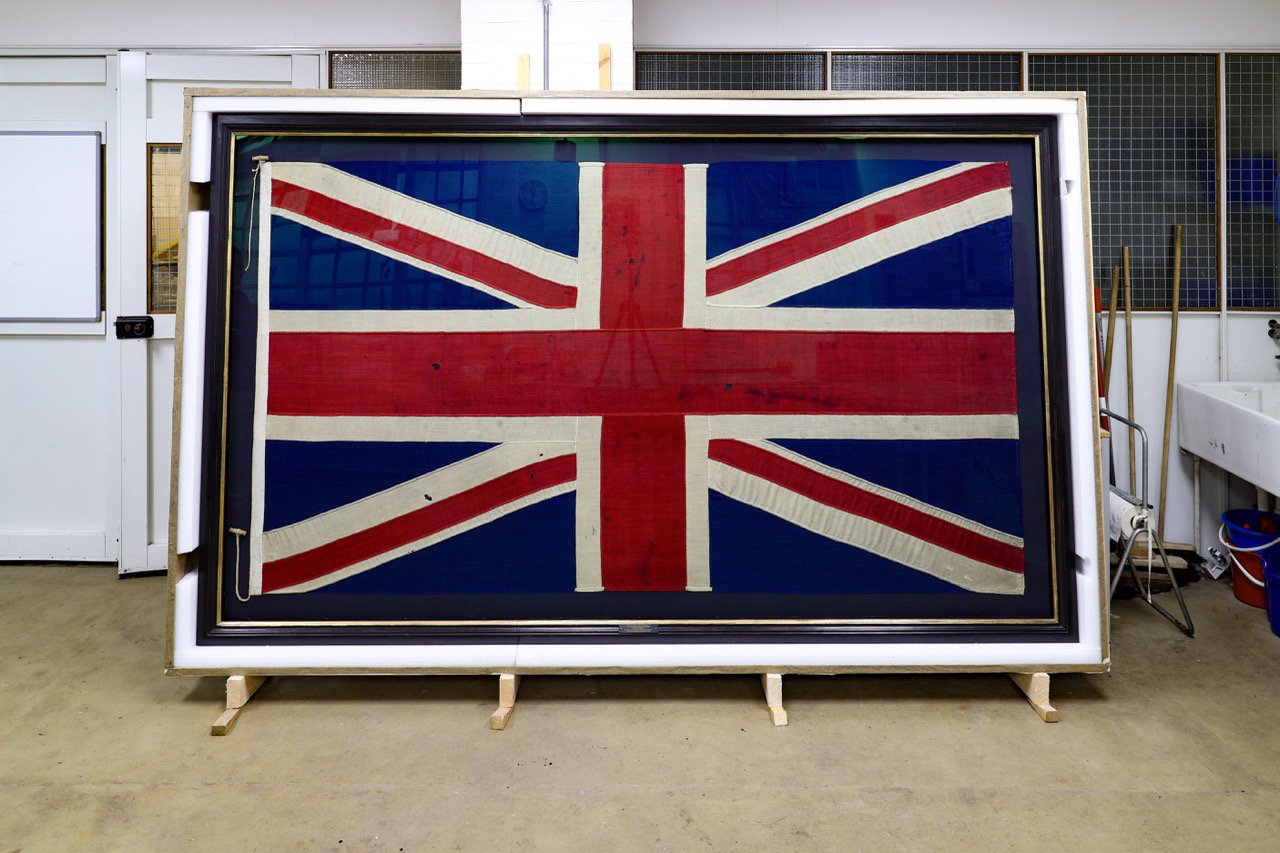A large Union flag, framed, sitting inside a wooden crate. A large Union flag, framed, sitting inside a wooden crate.