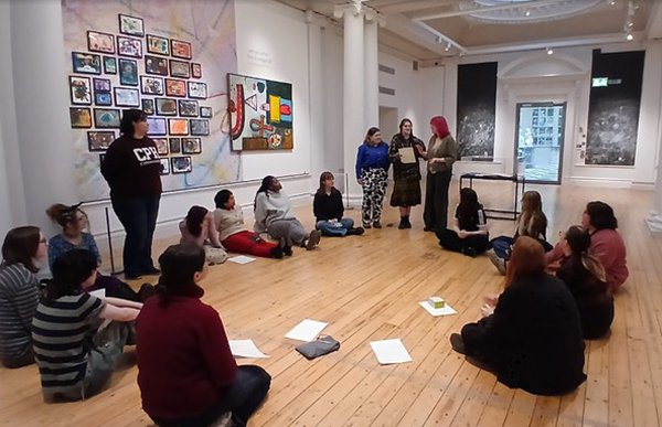 A group of young people sitting in a circle in a gallery. A group of young people sitting in a circle in a gallery.