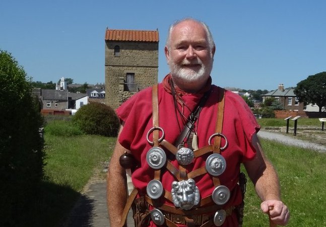 Man in a red tunic, standing with the West Gate in the background