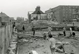 Black and white photograph of archaeologists watching the demolition of buildings while excavating a Roman fort site.