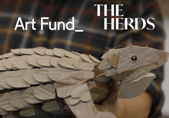 A photo of a cardboard Pangolin puppet. The image includes an ‘Art Fund’ logo and an ‘Inspired by the herds’ logo.