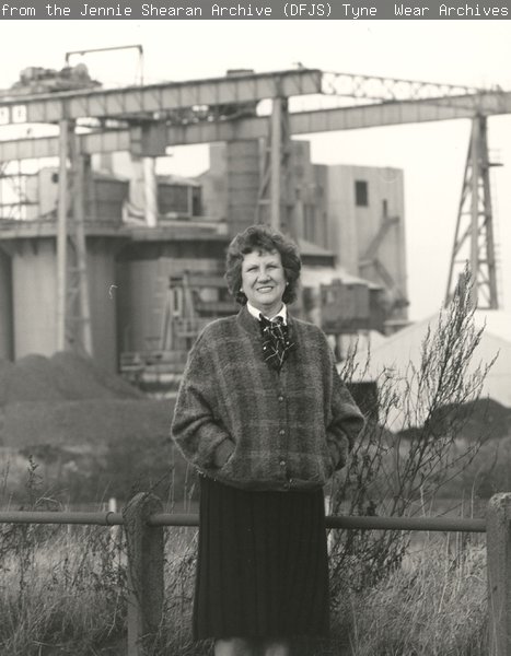 A black and white photograph of Jennie Shearan standing in a field with the coke works in the background.