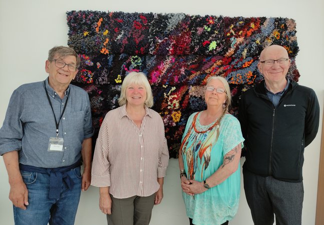 Four adults standing in front of a proggy mat on a wall