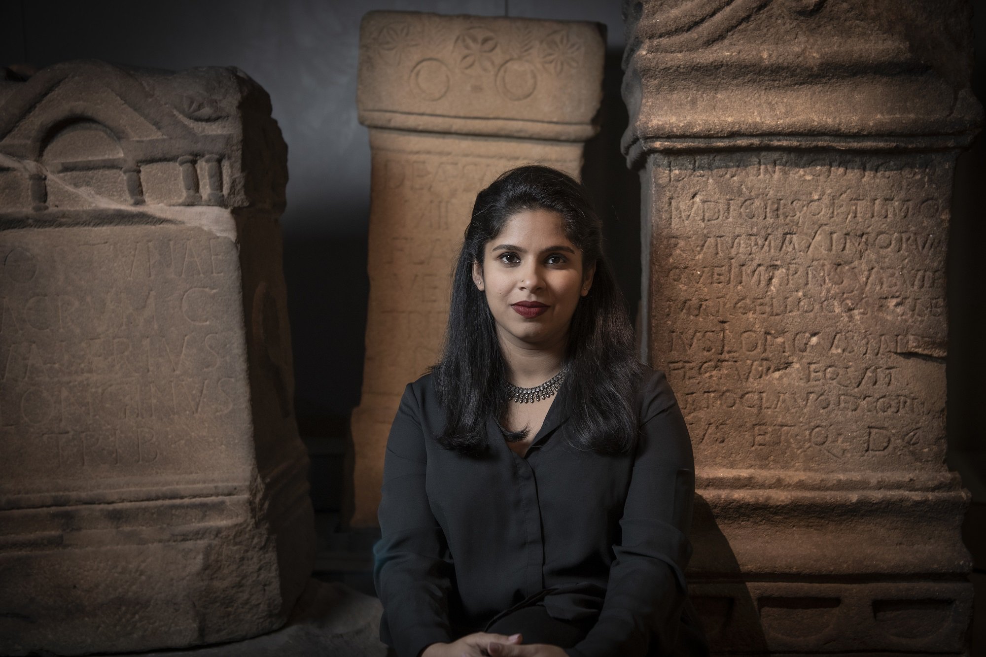 A woman in a black blouse sitting in front of sandstone Roman altars in a museum.