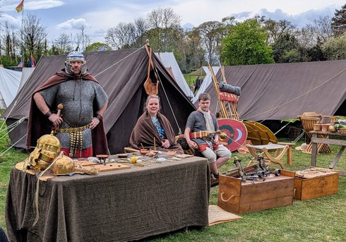 Historical reenactment camp with people in period costumes, tents, shields, helmets, and wooden tables with various items