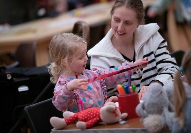 A photograph of a mum and her toddler crafting. The toddler holds a pink metallic ribbon.