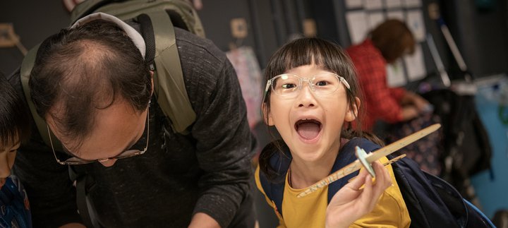 dad bends over table, daughter looks up holding out object she made