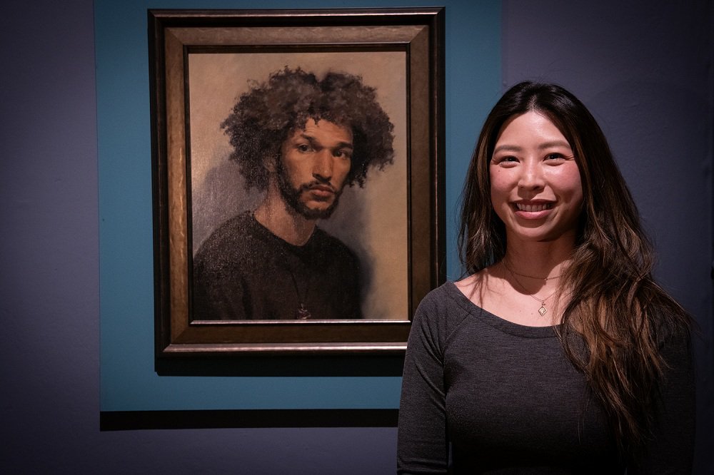 Woman standing next to her portrait painting of a man.