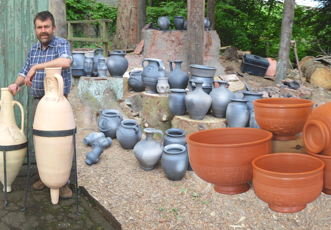 Man standing next to two amphorae beside a collection of different types of clay pots