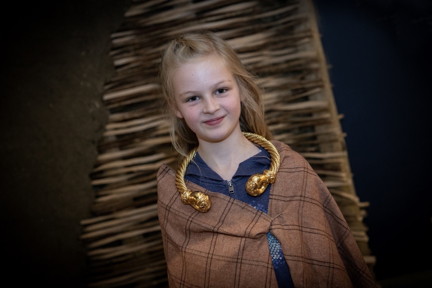 A child is dressed in Roman clothing and jewellery. They stand in front of a wicker backdrop