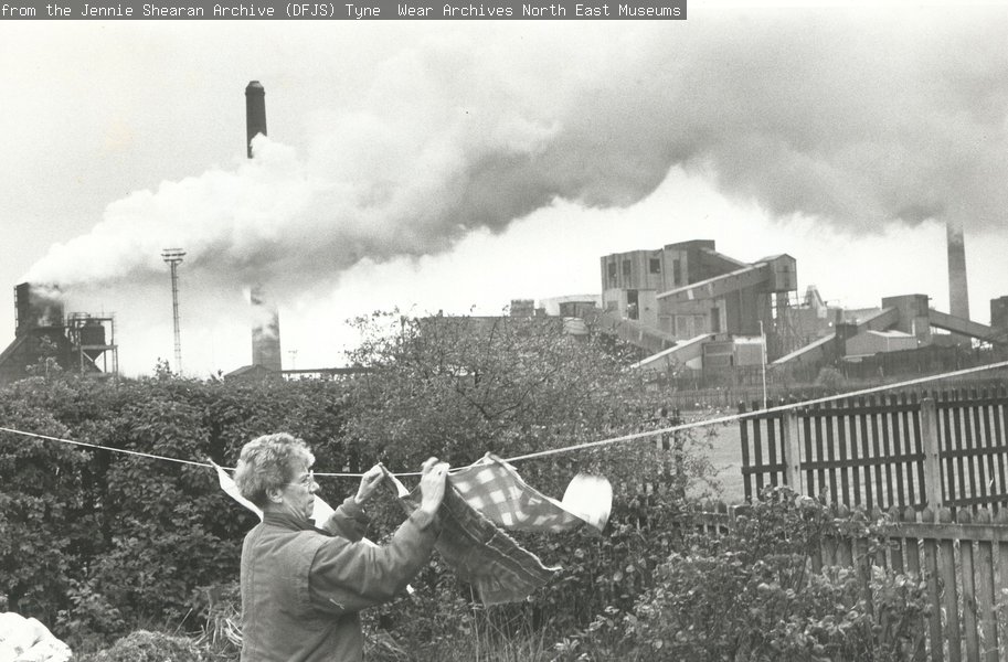 A black and white photograph of a woman hanging her washing out in her garden in front of a coke works with smoking chimney.