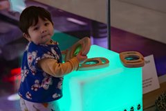 A toddler is standing next to light box cube which is placed on the ground, the toddler is holding a wooden toy.