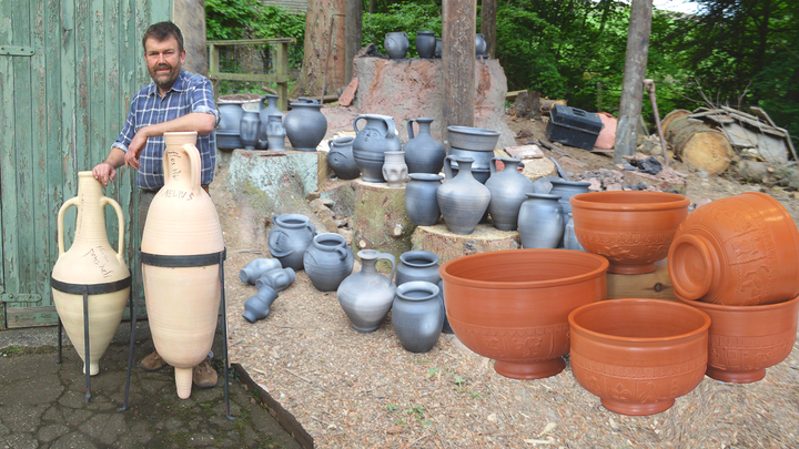 Man standing next to two amphorae beside a collection of different types of clay pots