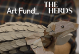 A photo of a cardboard Pangolin puppet. The image includes an ‘Art Fund’ logo and an ‘Inspired by the herds’ logo.