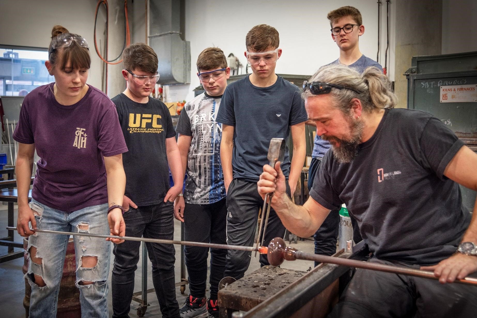 A group of students wearing eye protection gather around an artist who is working with glass A group of students wearing eye protection gather around an artist who is working with glass