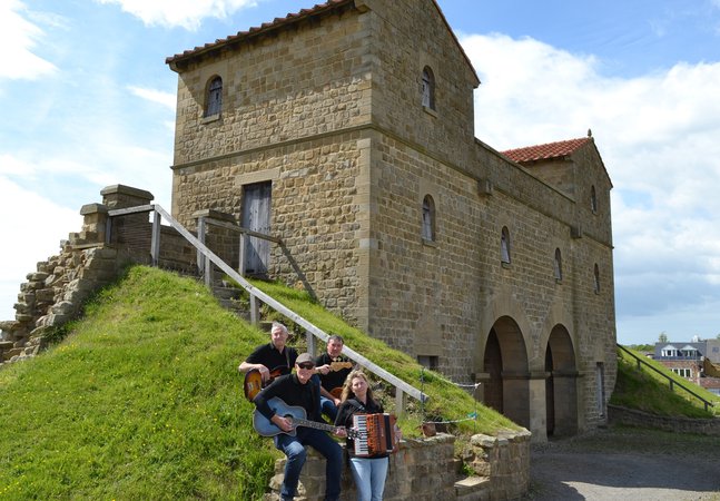 Four musicians sitting on a stone wall in front of a Roman style gatehouse