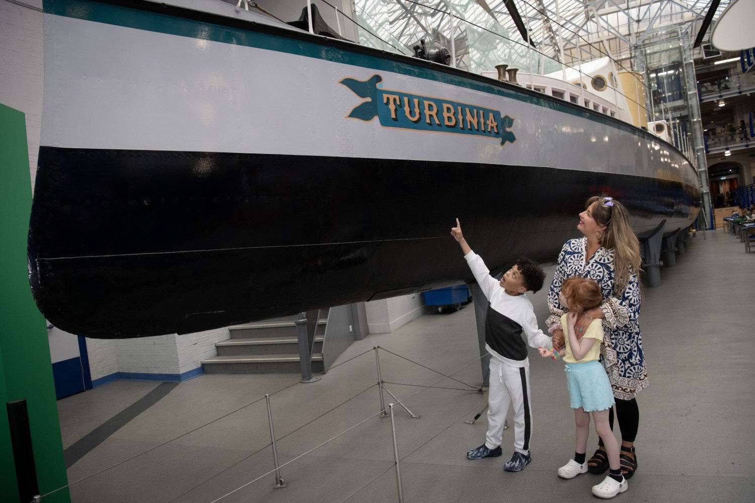 A family of three stand in Discovery Museum atrium and look up at the Turbinia ship.