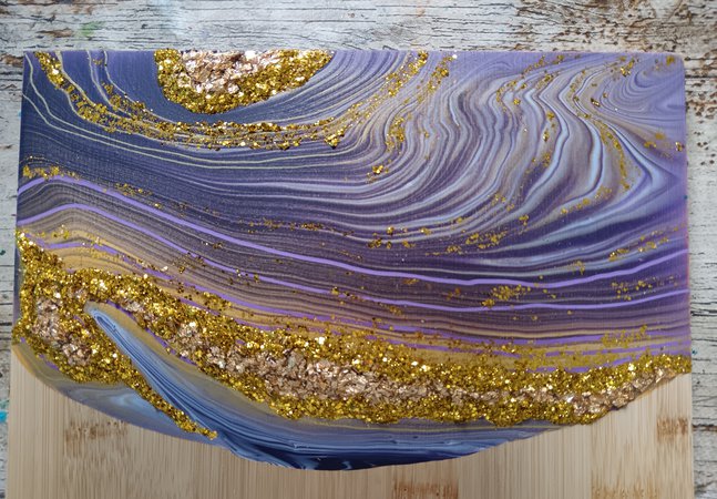 A photograph of an acrylic poured serving board. The decoration has purple swirls and gold decorations, inspired by geodes.