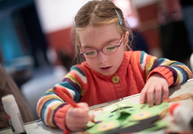 A photograph of a young girl colouring. She wears a striped jumper and glasses.