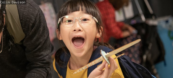 Girl i yellow top and glasses holds aeroplane with excited expression