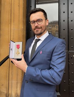 Smiling man with dark hair and glasses holds up an OBE medal