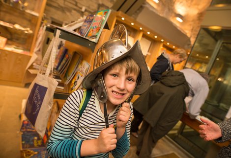 A child stands in a museum shop wearing a medieval helmet and smiling at the camera