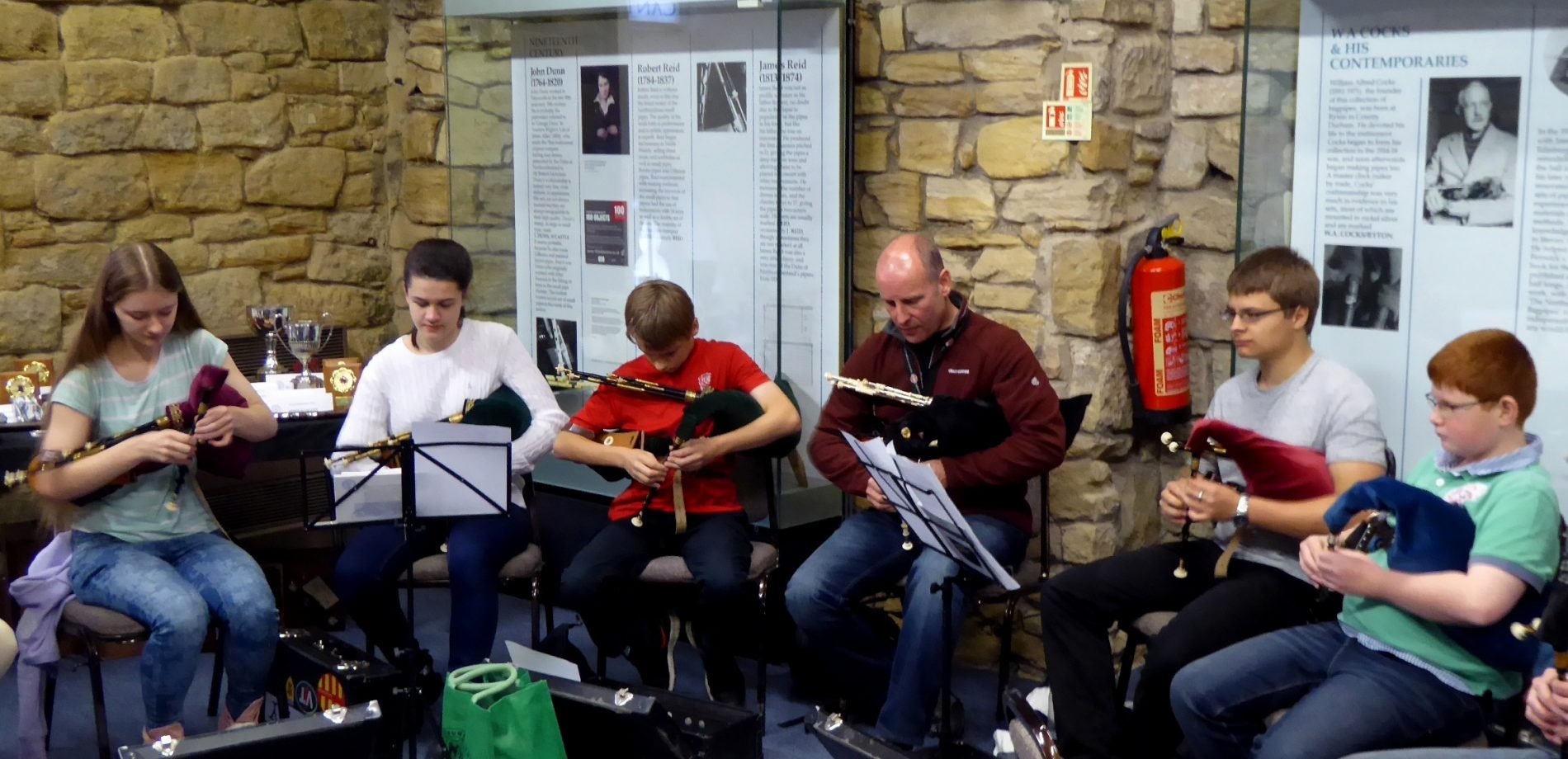 A group of pipers play bagpipes in a brick walled room with display panels behind them.