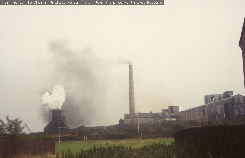 A photograph of a house estate in the foreground and a child riding his bike, and a coke works with black smoking chimneys in the near background.