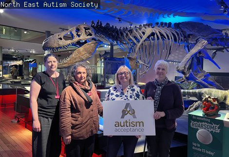 hotograph of 3 people in front of a T-Rex skeleton in the Fossil Stories Gallery at Great North Museum: Hancock being presented with a white board reading Autism Acceptance by another person.