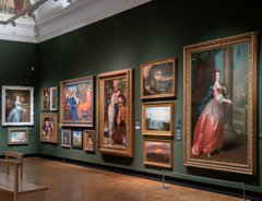A photograph of the inside of the gallery. Oil paintings in frames hang on a green wall.