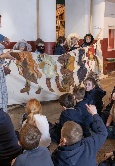 Person dressed as an ancient Brit talking to a group of schoolchildren sitting on the floor with a cloth timeline of clothing throughout the ages 