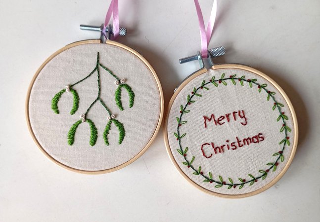 Two embroidery rings with cream fabric. One has a green and white mistletoe design and the other reads 'Merry Christmas' in red thread with a green wreath border