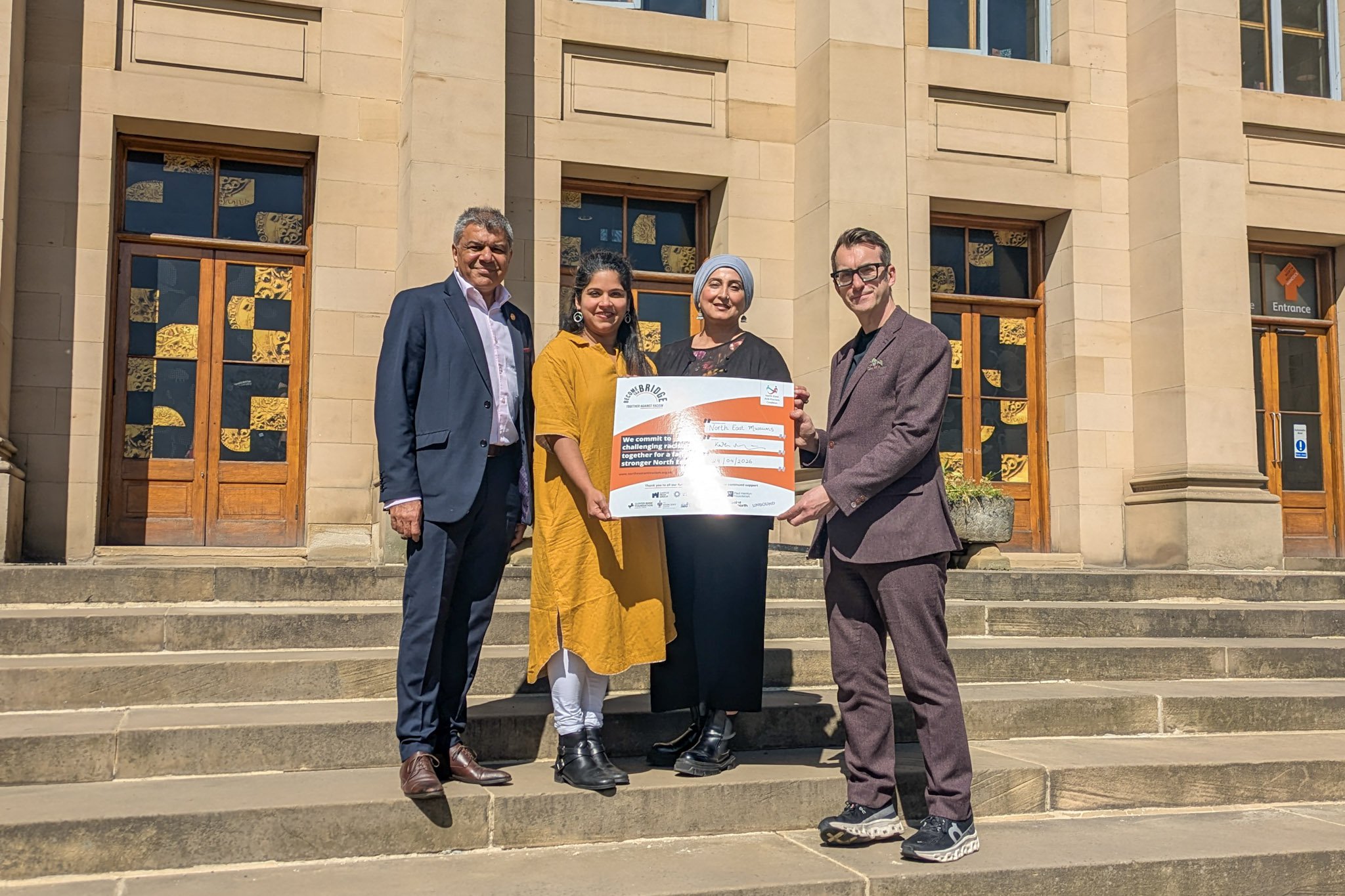 Four people holding an oversized pledge card outside a museum