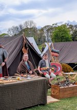 Historical reenactment camp with people in period costumes, tents, shields, helmets, and wooden tables with various items