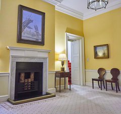 A room with a marble fireplace with a painting of a bridge hung over it. Walls are painted yellow.