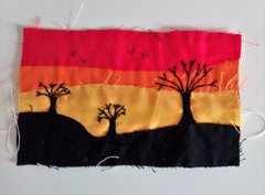 An embroidered landscape with trees silhouetted against a red, orange, and yellow sky. 