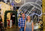 A man in a checked shirt and a woman in a patterned dress standing in front of large Christmas decorations with lots of twinkly fairy lights