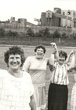 Black-and-white historical photograph of six community members standing on a grassy field in front of an industrial site. A brown haired woman in the foreground holds a document up.