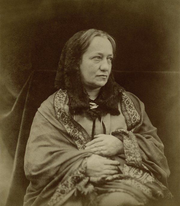 A sepia photograph of a seated woman. She is wearing a dark hood and a shawl with a paisley design border.