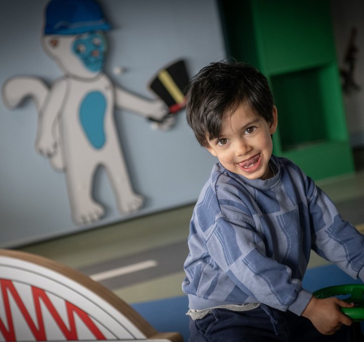 A small child looks at the camera, tongue sticking out. They are sat down, holding on to a toy, plastic steering wheel. In the background is a model of a bridge and an illustration of a cat holding a hat