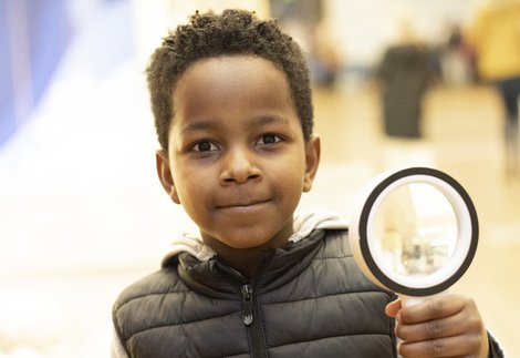 A boy looks into the camera, holding up a magnifying glass
