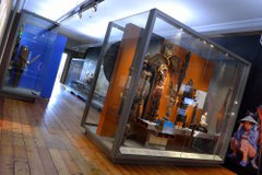 View of ethnographic items in glass museum cases. There are headdresses, statuettes and suits of armour