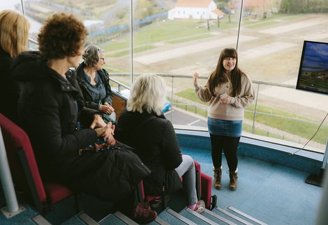 A person with long brown hair giving a talk to a small group of people.