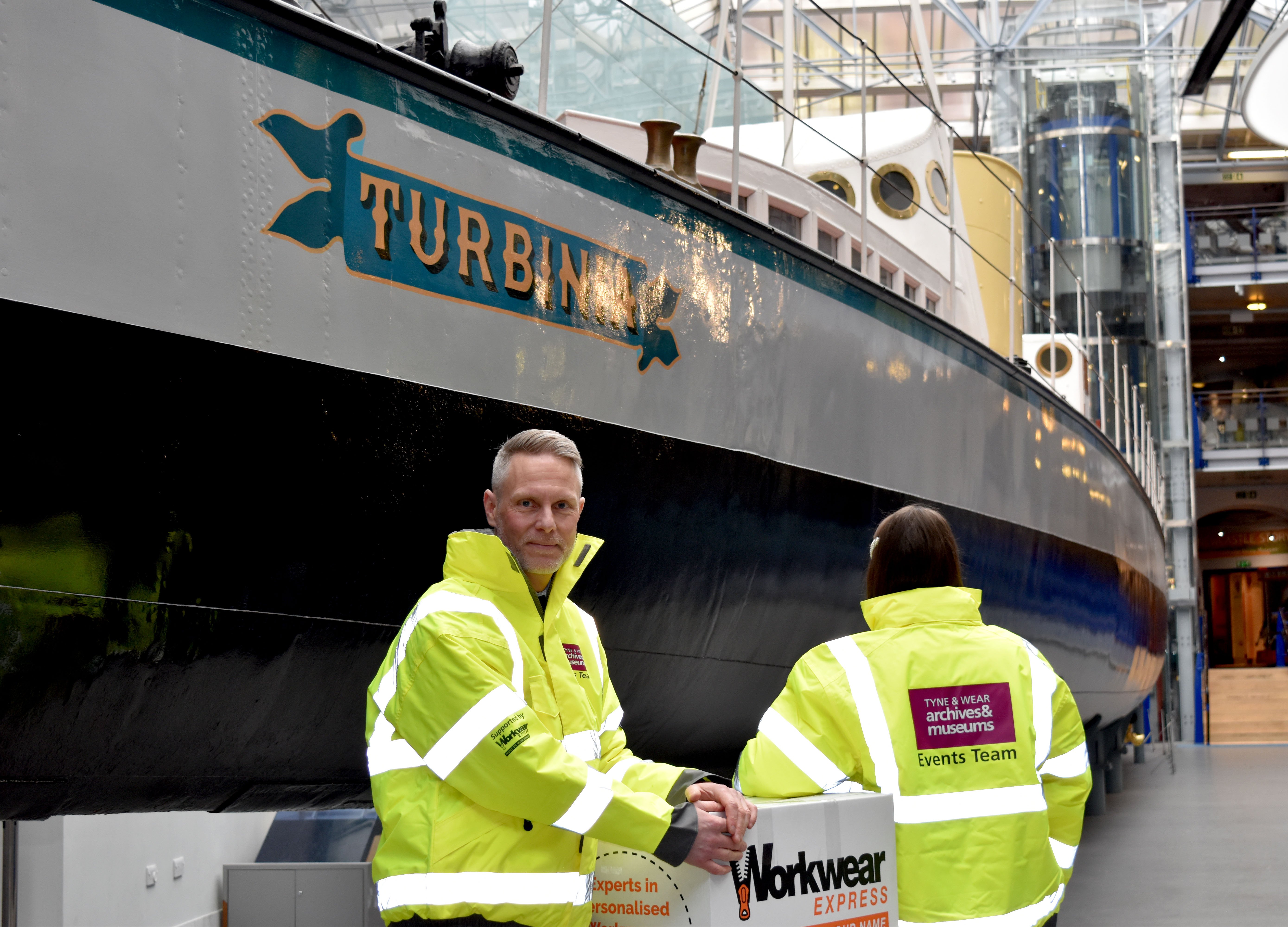 A man and woman wear high visibility yellow jackets beside the Turbinia exhibit in Discovery Museum, Newcastle