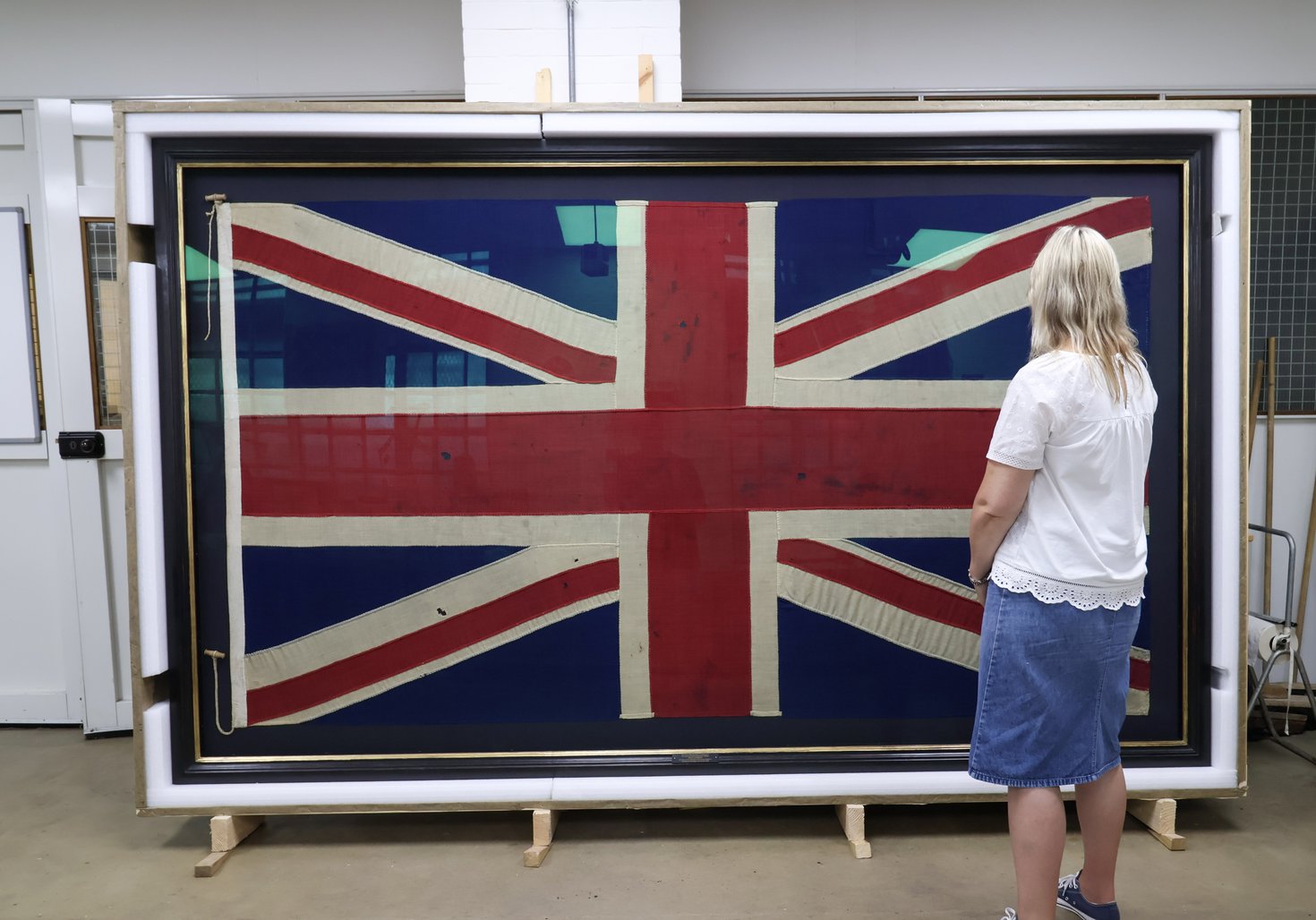 A large framed Union Flag in behind glass with a blond woman looking at it