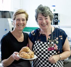 Two women smiling, holding a plate of scones with kitchen equipment in the background