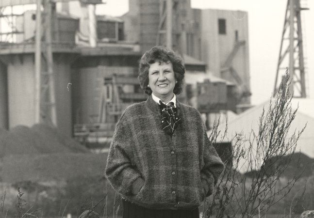 black and white photo of smiling woman with short brown curly hair standing in front of what appears to be an industrial site