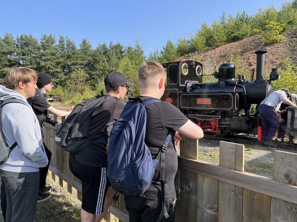 A group of teenagers watching staff member working by steam train